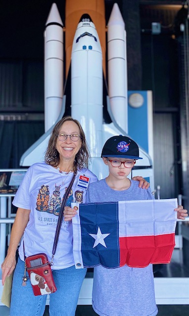 a young student and his mother standing side by side in front of a large space shuttle replica. The boy is holding a Texas flag