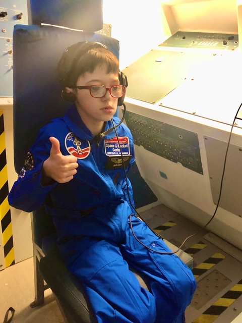 a boy sitting in the captain's chair of a mock space shuttle, giving a thumbs up