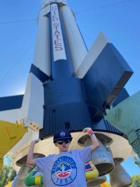 A young male student flexing his muscles in space camp t shirt, hat and sunglasses, standing in front of a giant United States rocket statue