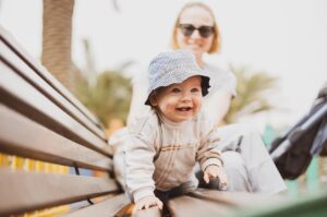 A 1 year old in a fishing hat crawls across a park bench, in front of mother