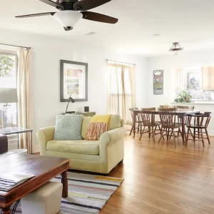 A picture of a big sunlit room with hard wood floors, big comfy couches, an inviting kitchen and dining area, and big windows