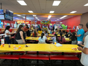 Students and teachers sit at colorful tables in a pizza place and arcade