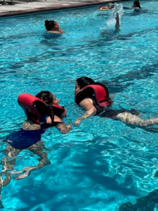 Students swim in a pool with bright red life vests on.