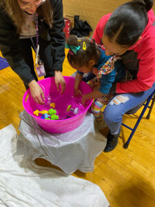 A Young girl reaches into a colorful bucket for toy frogs with two smiling women in a gymnasium