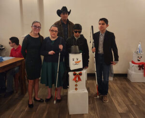 A group photo of children with visual impairments and their father with a snowman made out of cardboard boxes and white&black wrapping paper at the region 1 winter gala, December 2023