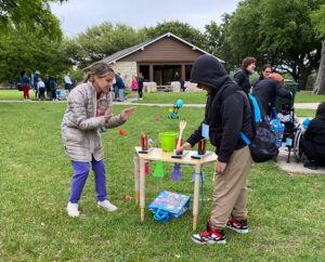 A boy in a hoodie and an older woman play with a spoon catapult on a table with bunny decorations. They are on the lawn in front of a pavilion