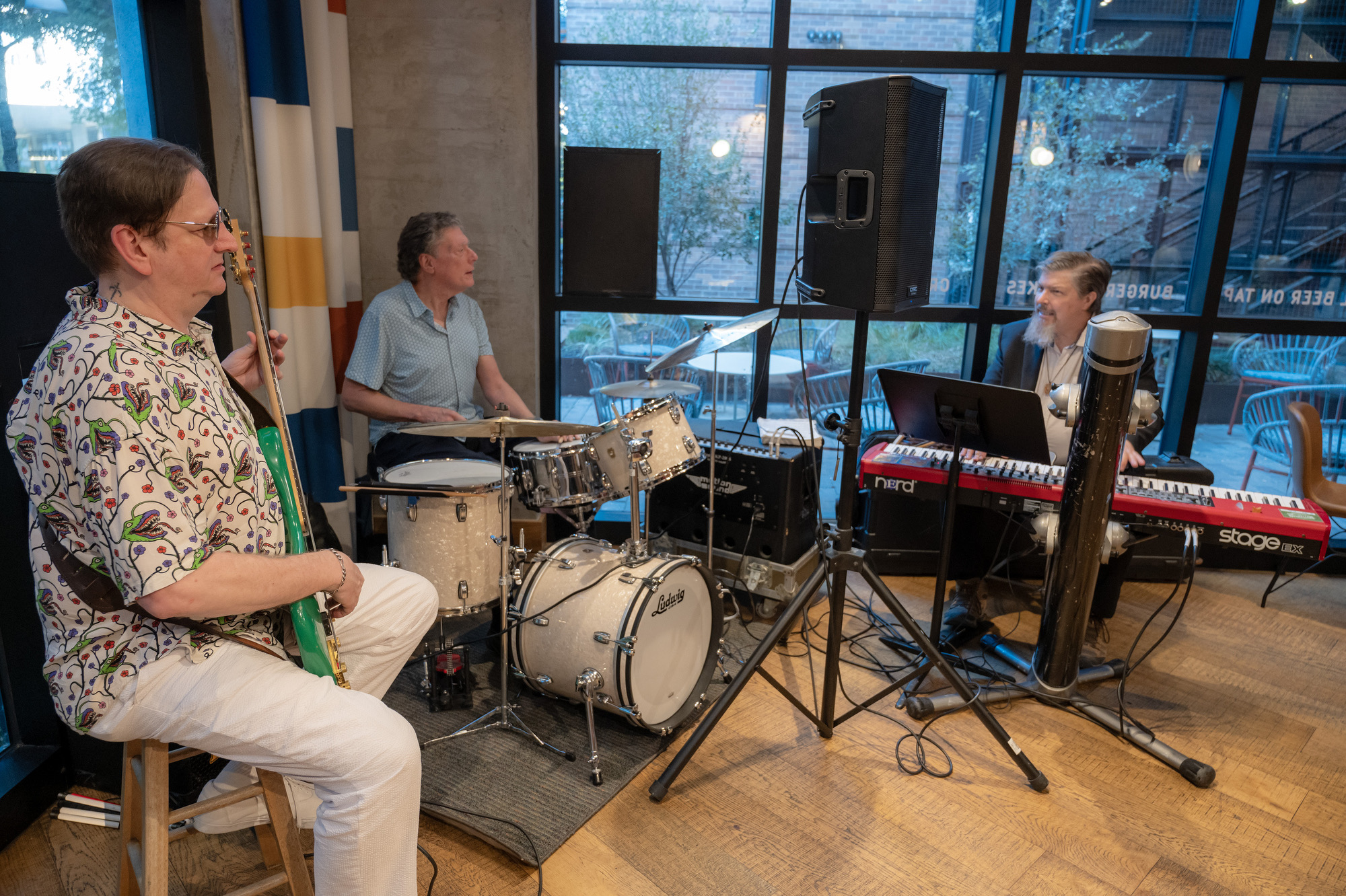 Three musicians perform together indoors, with one playing guitar, one on drums, and one on a keyboard near large windows.