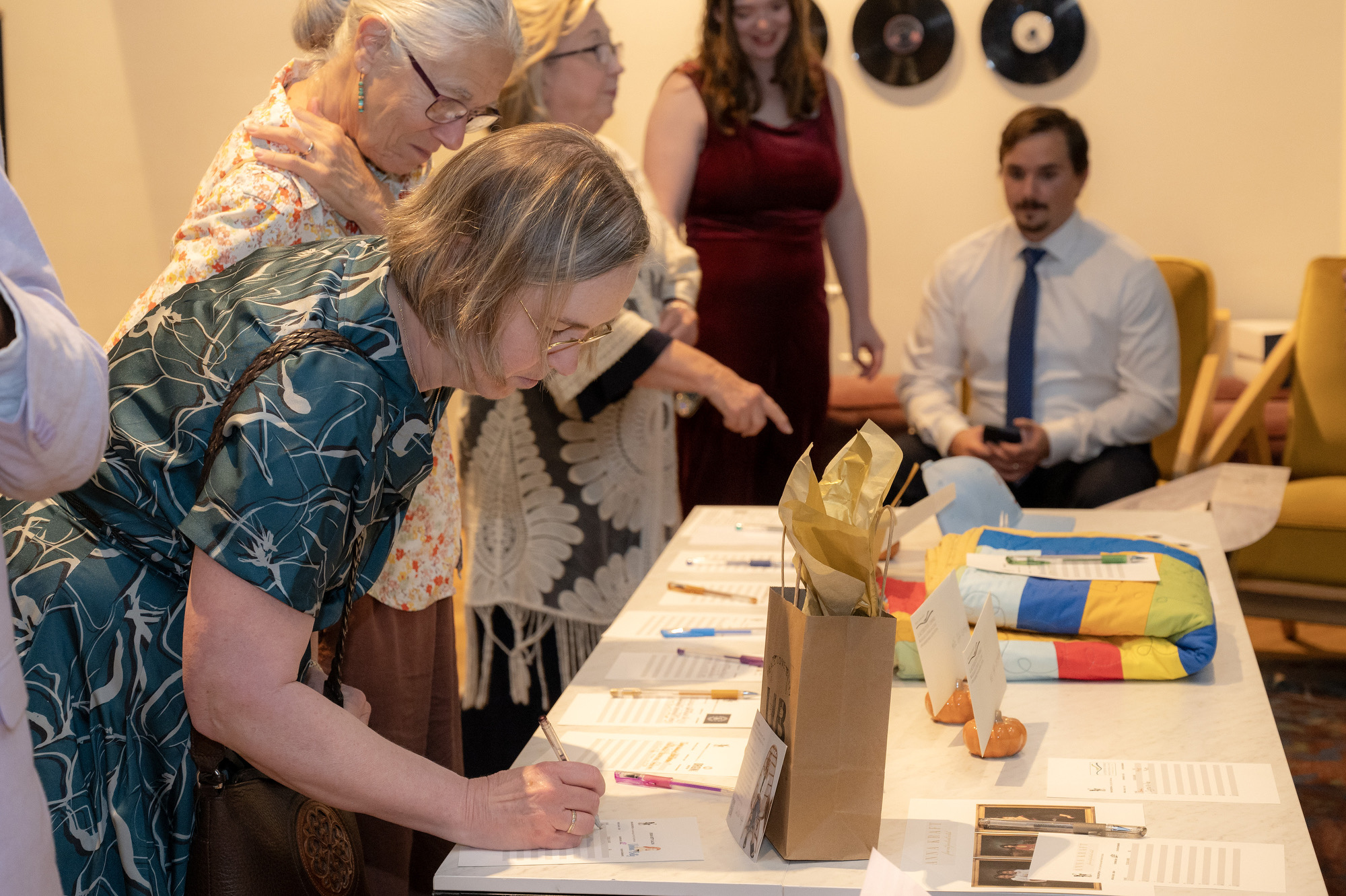 A woman signs a form at a table during a community event while others stand nearby. The table displays papers, pens, and colorful items, suggesting a registration or silent auction setting.