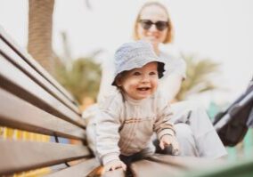 A 1 year old in a fishing hat crawls across a park bench, in front of mother